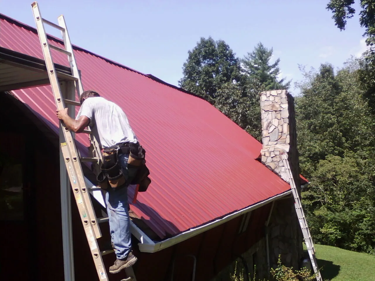 Roofer on a ladder beside a red metal roof and stone chimney — Soffit & Fascia Repair by Roofing Brothers in Garden Plain